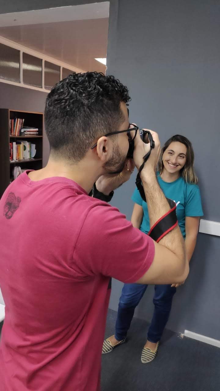 Homem de camiseta vermelha fotografa uma mulher sorridente de camiseta verde-água encostada numa parede cinza de escritório